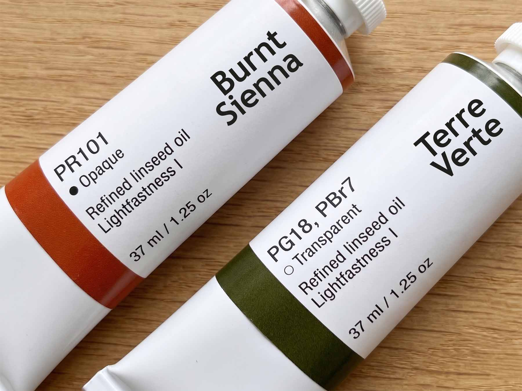 Two artist oil paint tubes on a wood surface, one labeled "Burnt Sienna / PR101" and one labeled "Terre Verte / PG18, PBr7," showing the Colour Index code printed beneath the color name on each label.